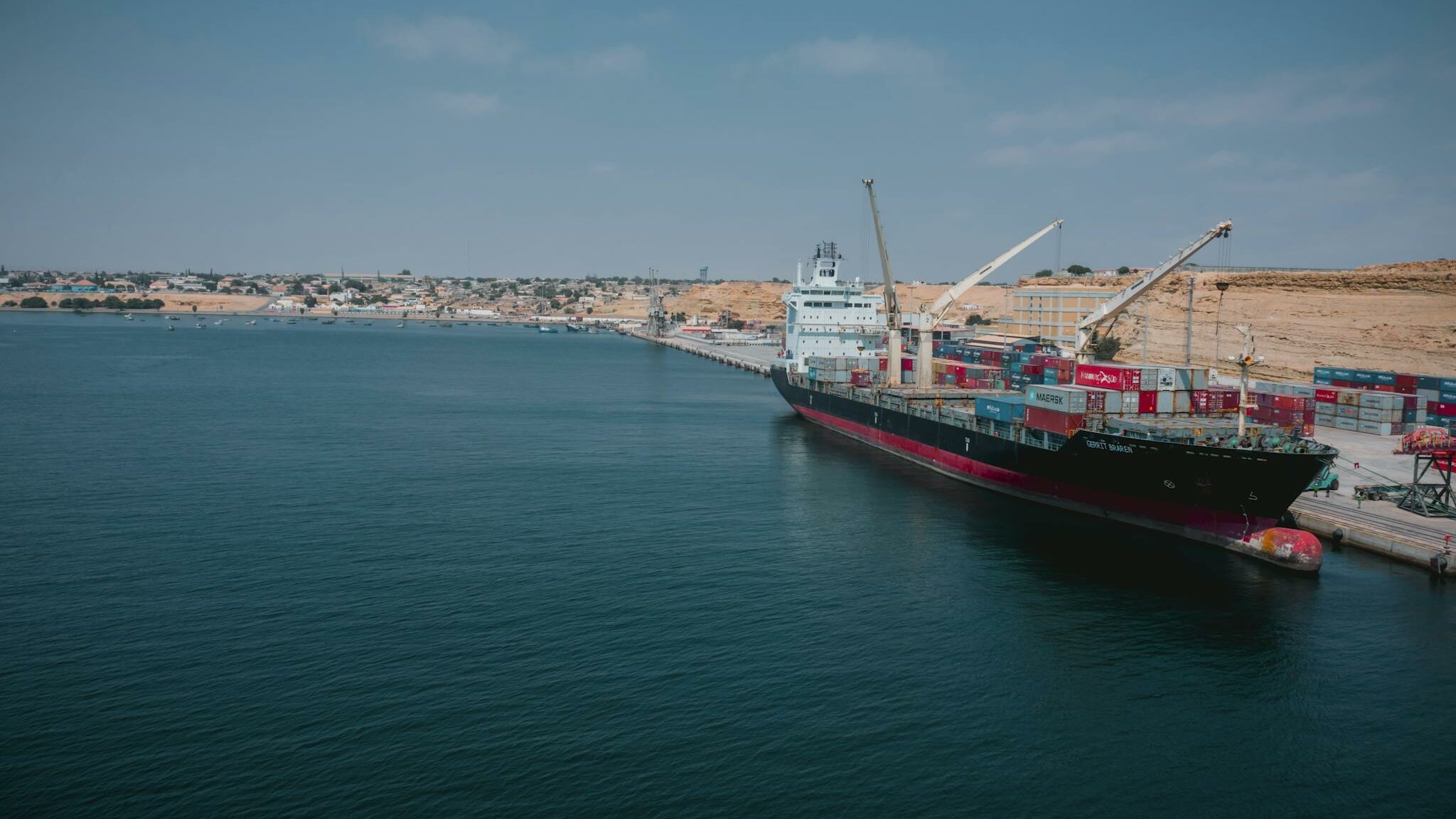 Cargo ship docked at Namibe port in Angola with shipping containers and cranes.